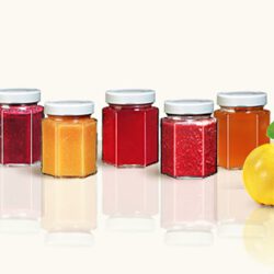 5 Sechseck-Twistoffgläser mit 3 unterschiedlichen roten und zwei orangefarbenen Marmeladen. Sie spiegeln sich zart im glänzend weißen Untergrund. Eine halbe leuchtend gelbe Quitte ist am rechten Bildrand zu sehen.